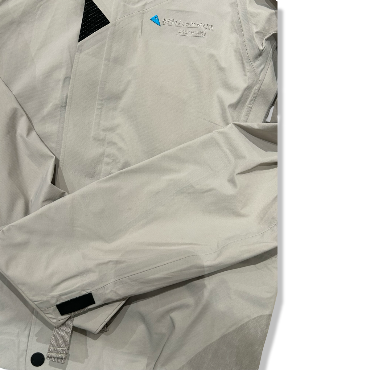 Close-up of a beige and gray Klättermusen down jacket with an embroidered logo on the chest.