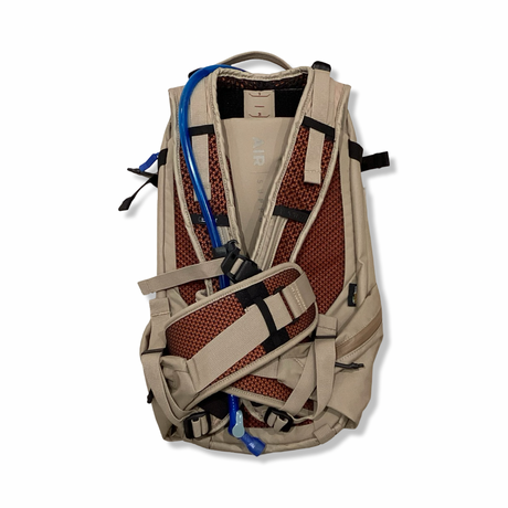 Baksidan av en beige Camelbak HAWG 20 L ryggsäck med vadderat bärsystem och blå vätskeslang, mot vit bakgrund.