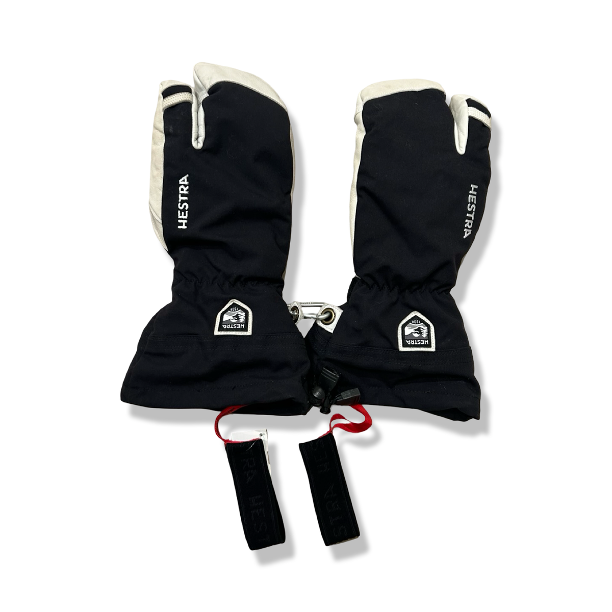 A pair of black and white Hestra three-finger gloves with wrist straps, front view against a white background.