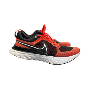 Orange och svarta Nike React löparskor för herr med vit sula, sedda från sidan mot vit bakgrund.