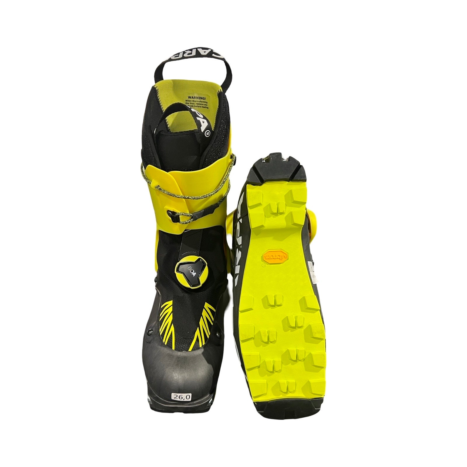 Scarpa Alien Carbon Grimalid pjäxor för herr i svart och gult, fotograferade mot en vit bakgrund. En pjäxa visas framifrån och den andra visar sulan.