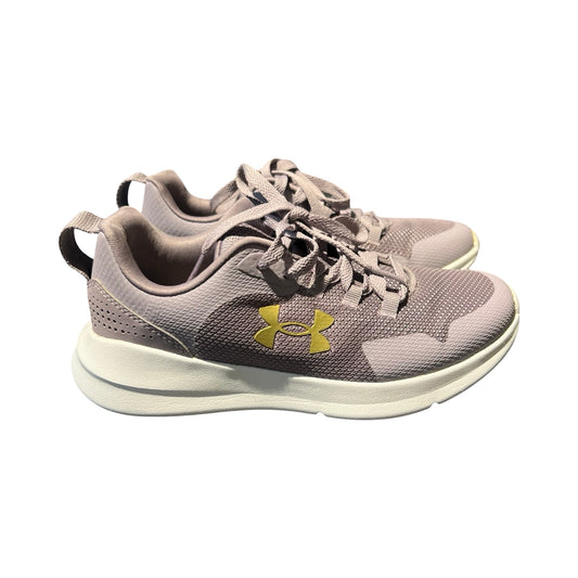 Ett par Under Armour träningsskor för dam i ljuslila med vita sulor och en guldfärgad logotyp. Skorna är fotograferade från sidan mot en vit bakgrund.